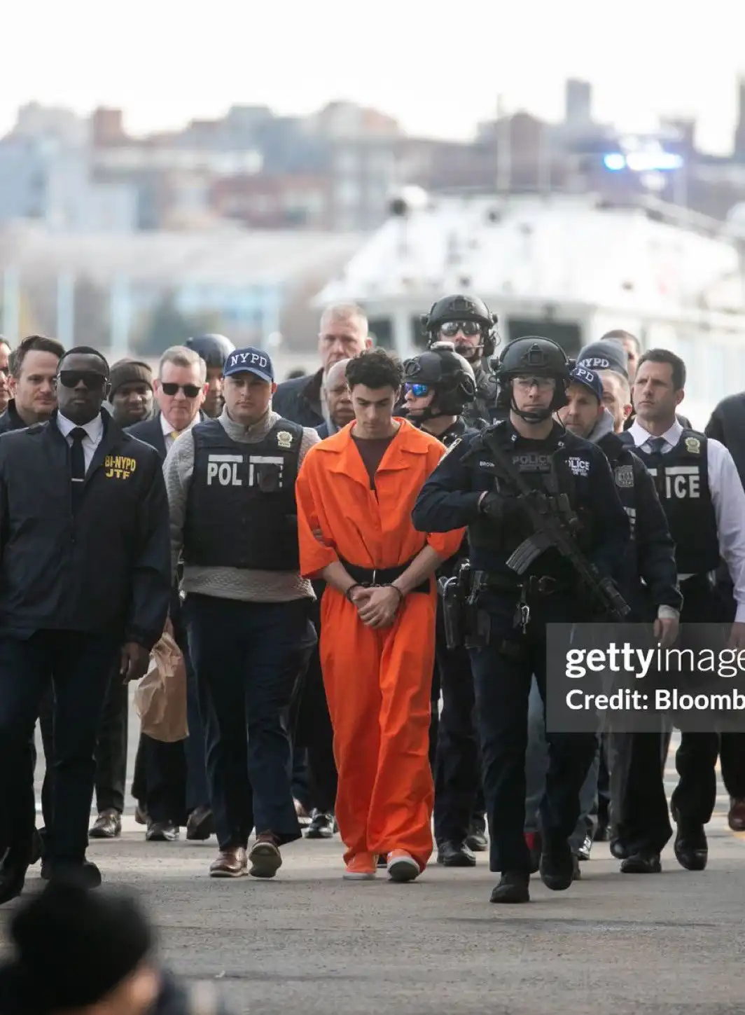 A young man in an orange jumpsuit is being escorted by numerous police officers, including those with 'NYPD' and 'FBI-NYPD…