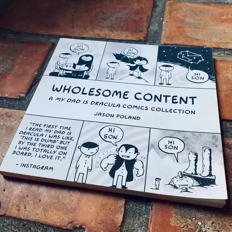 A comic book titled 'Wholesome Content: A My Dad Is Dracula Comics Collection' by Jason Poland rests on brick pavement. The…