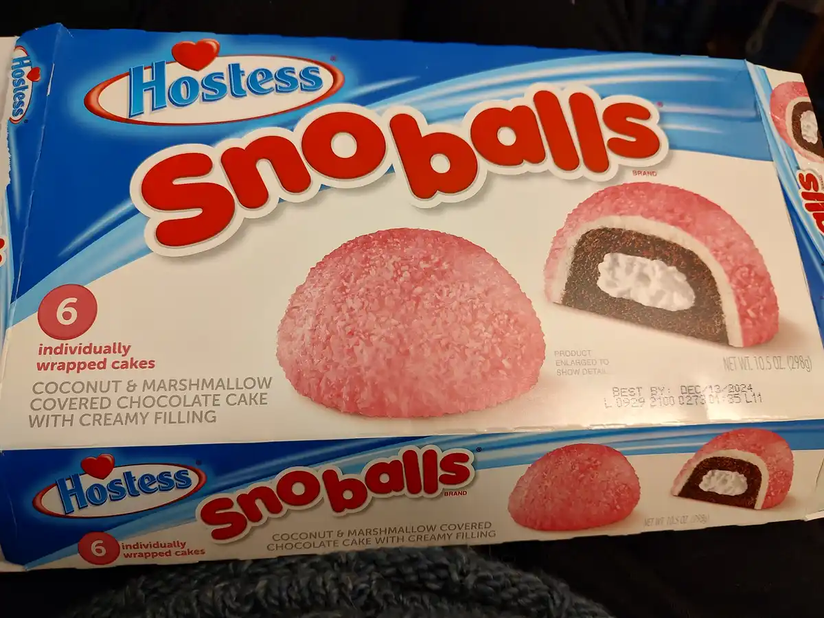 A close-up of a Hostess Snoballs snack cake box, showing pink coconut-covered chocolate cakes with creamy filling. The…