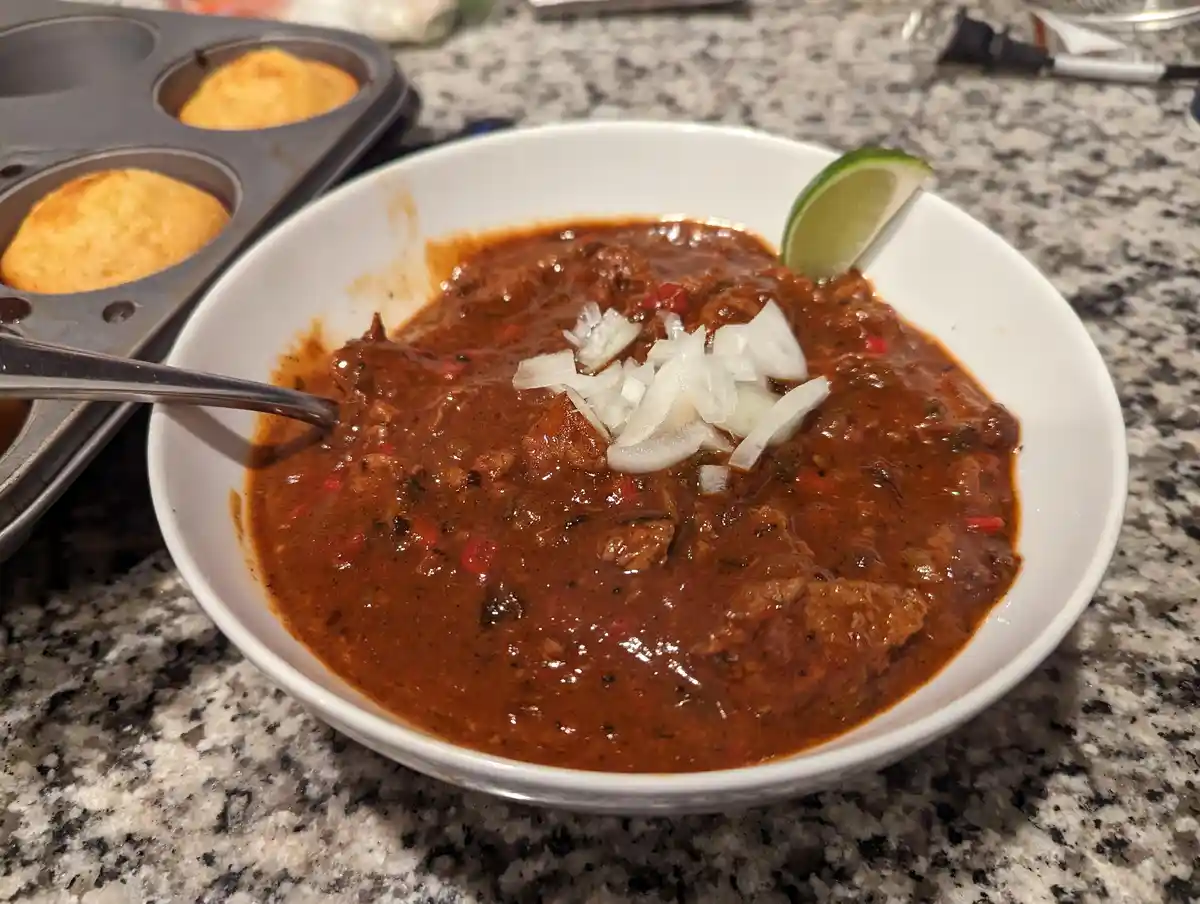 A close-up photo shows a white bowl filled with thick, dark red chili topped with chopped white onions and a lime wedge on…