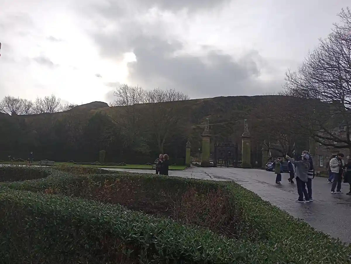An overcast day at a historic estate with manicured hedges in the foreground, a grand gated entrance, and bare trees…