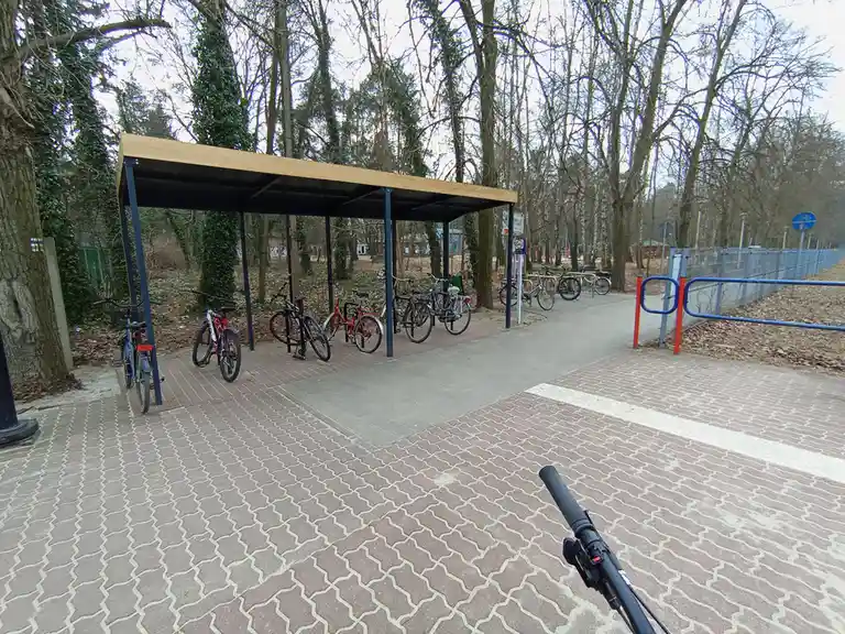 A first-person view from a bicycle, looking at an outdoor bike rack with several bikes parked under a wooden roof…