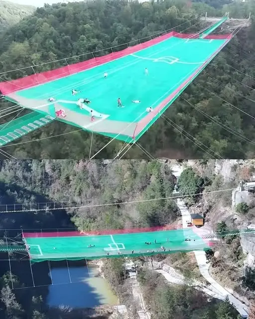 An aerial view shows a green and red football field suspended high over a valley, with people playing soccer on it. The…