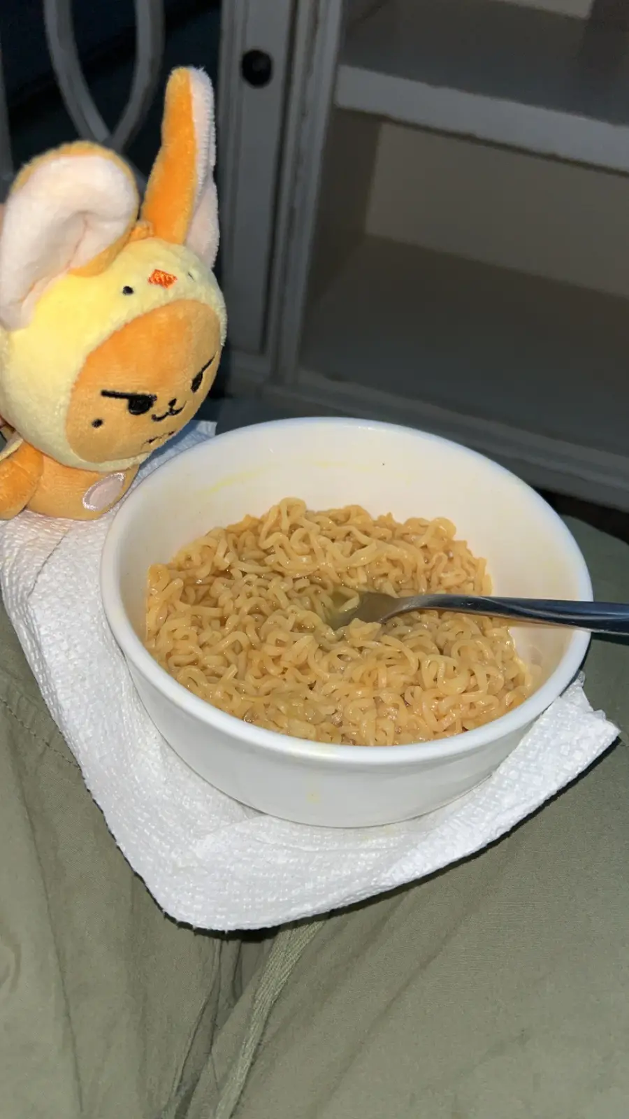 A close-up photo shows a white bowl of instant ramen noodles with a fork resting in it, placed on a white paper towel on…