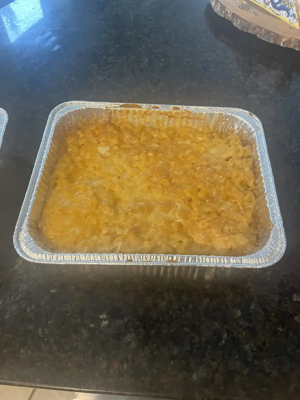 A rectangular aluminum foil baking dish filled with golden-brown, bubbly macaroni and cheese sits on a dark speckled…
