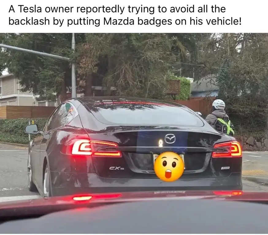 A black Tesla Model S is shown from behind, with a Mazda logo sticker placed over its actual Tesla emblem on the trunk. The…