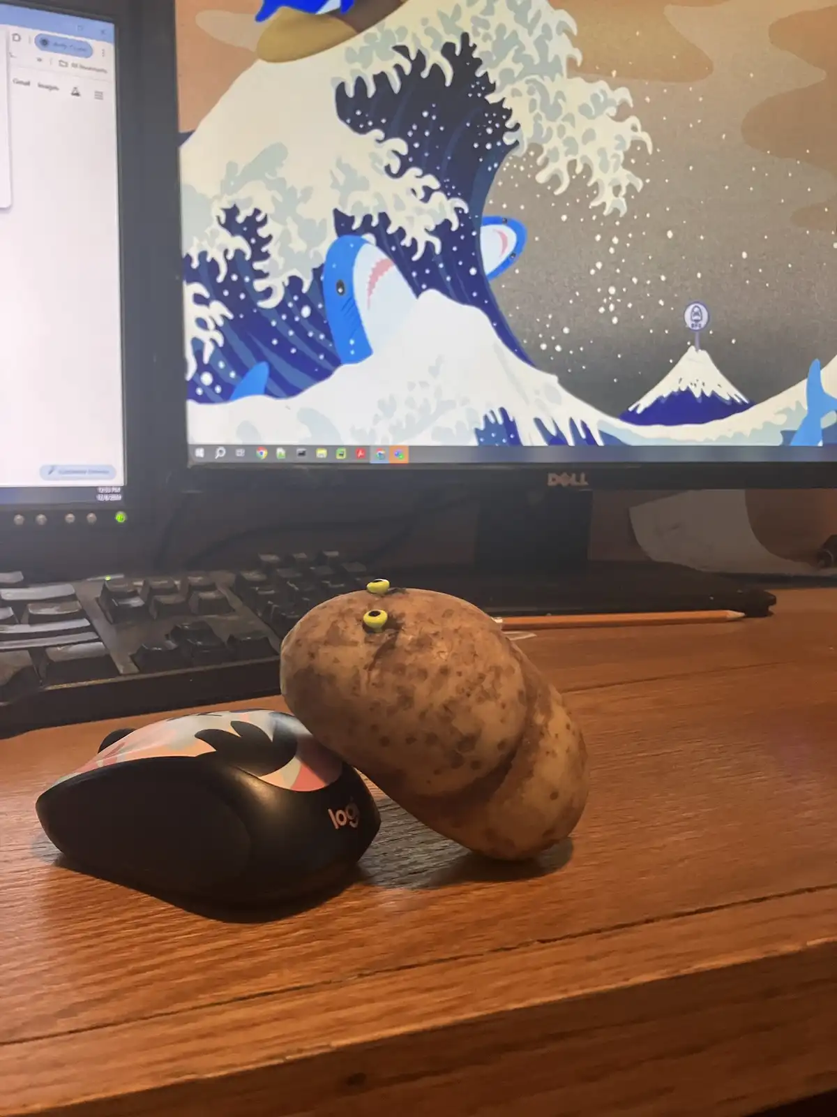 A potato with two small yellow googly eyes is leaning against a Logitech mouse on a wooden desk, in front of a Dell monitor…