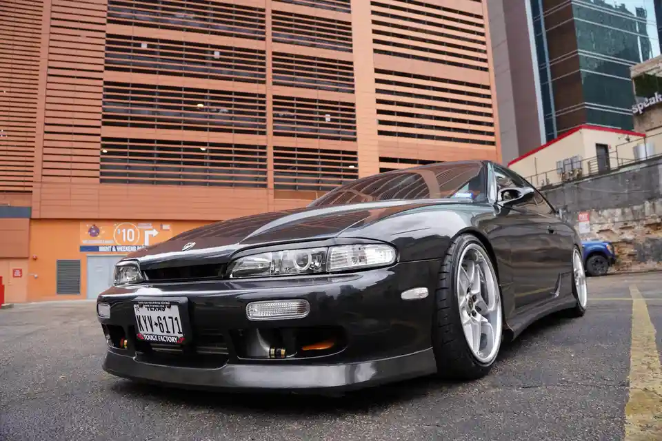 A black Nissan Skyline R34 sports car with custom silver wheels and a lowered stance is parked in an urban parking lot in…