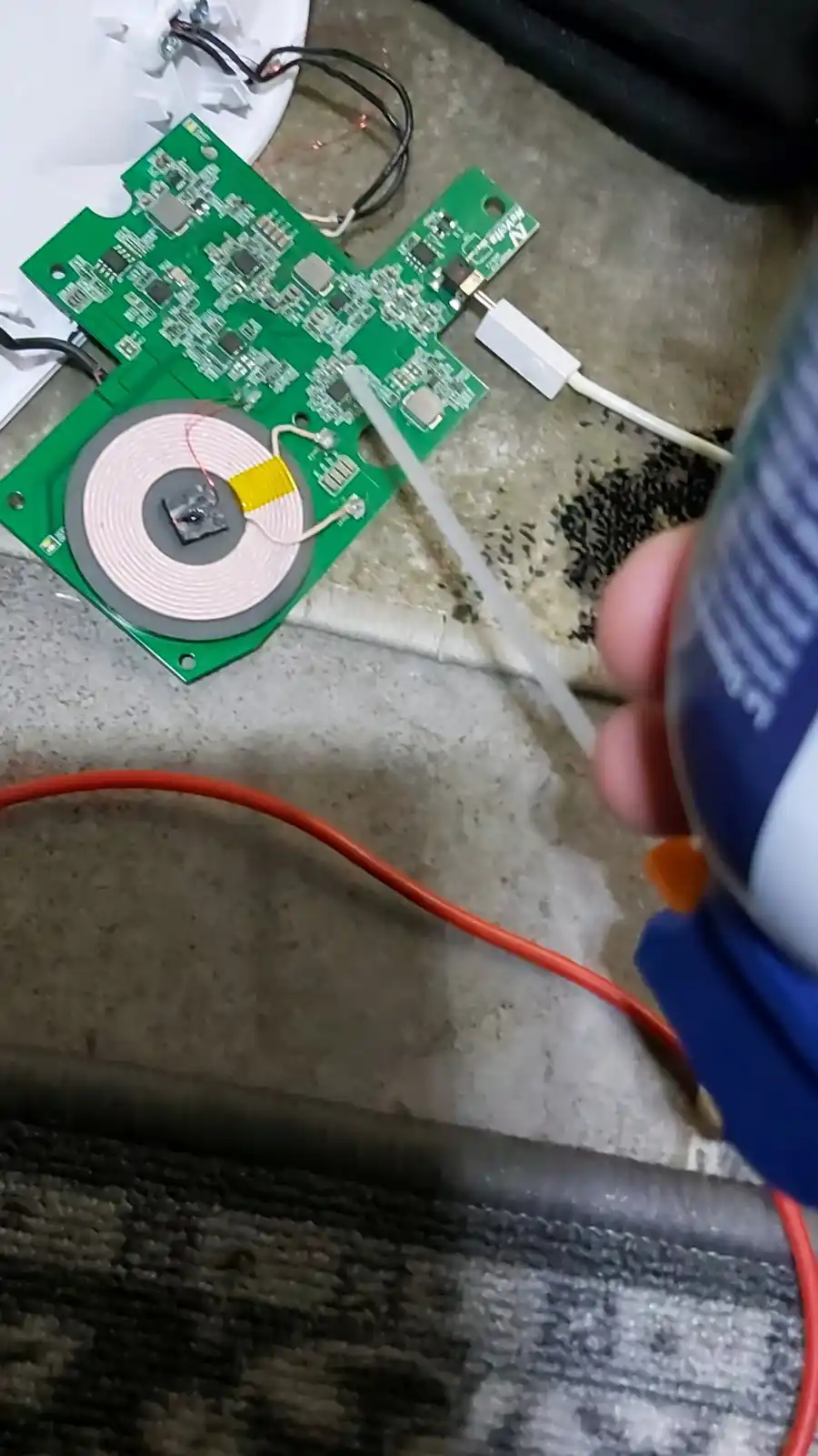 A close-up photo shows a hand holding a can of WD-40 spray, aiming it at a green circuit board lying on a concrete floor…