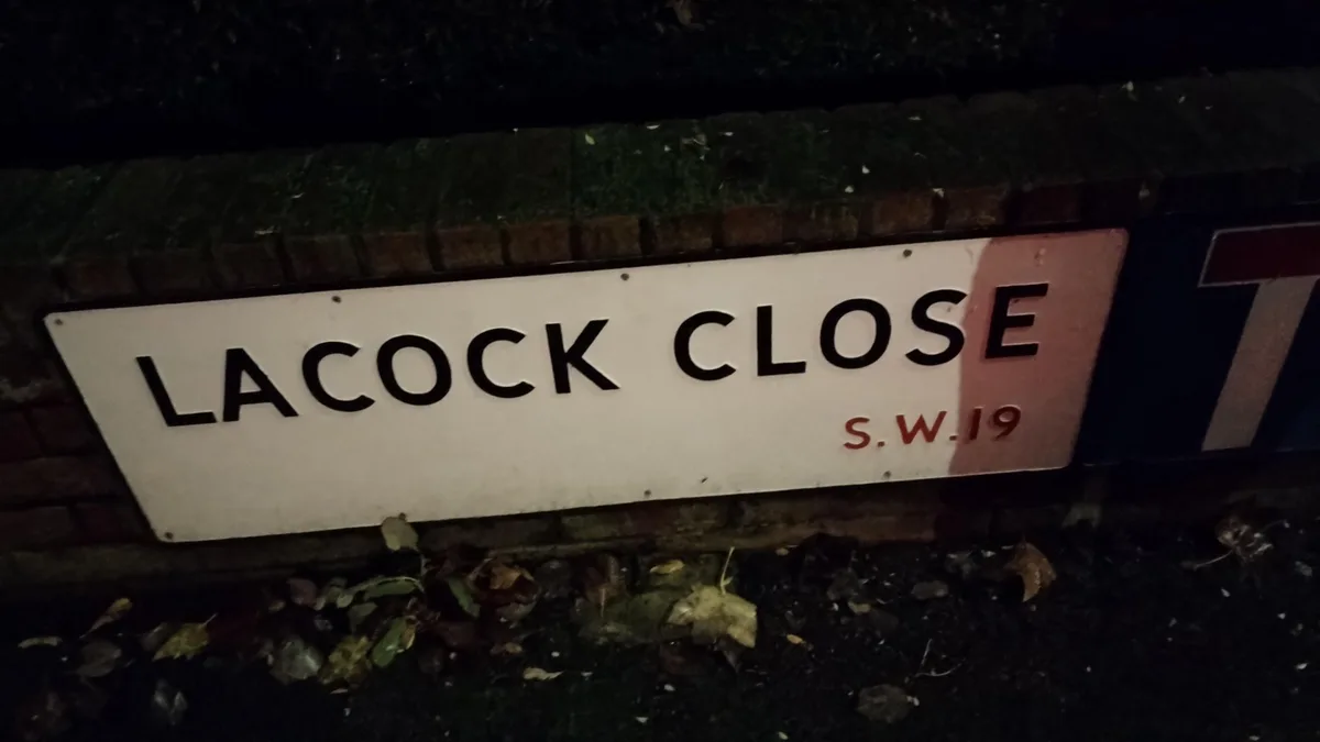 A white street sign reading 'LACOCK CLOSE' with 'S.W.19' below it, mounted on a brick wall at night, partially obscured by a…