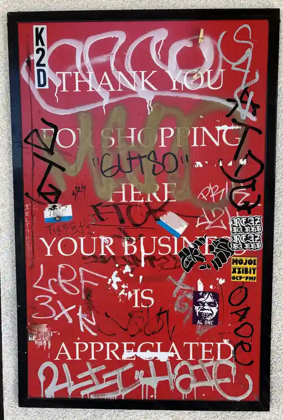 A red, framed sign covered in layers of graffiti and stickers, with the original text 'THANK YOU FOR SHOPPING "GUTSO" HERE…
