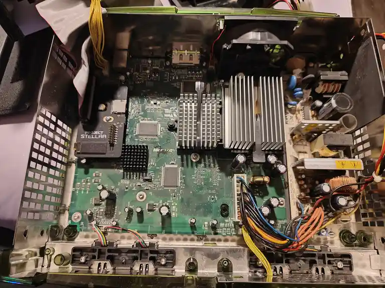 A close-up photo of the internal circuit board of a Microsoft Xbox console, showing its green motherboard, heat sinks…