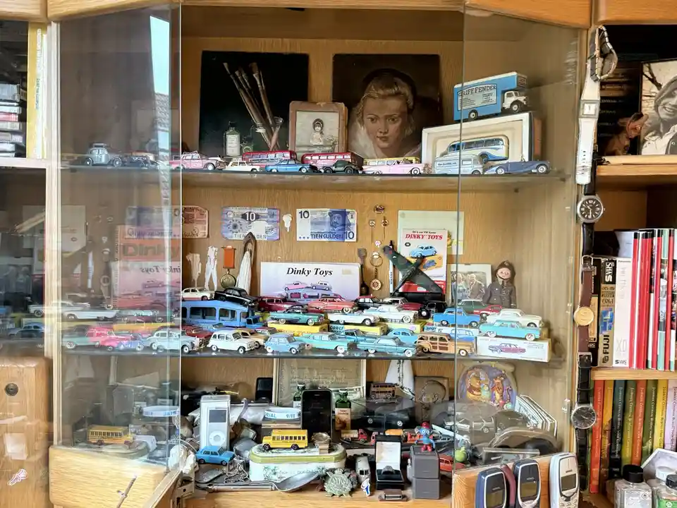 A glass-fronted wooden display cabinet is densely packed with vintage Dinky Toys die-cast vehicles, old Dutch banknotes…