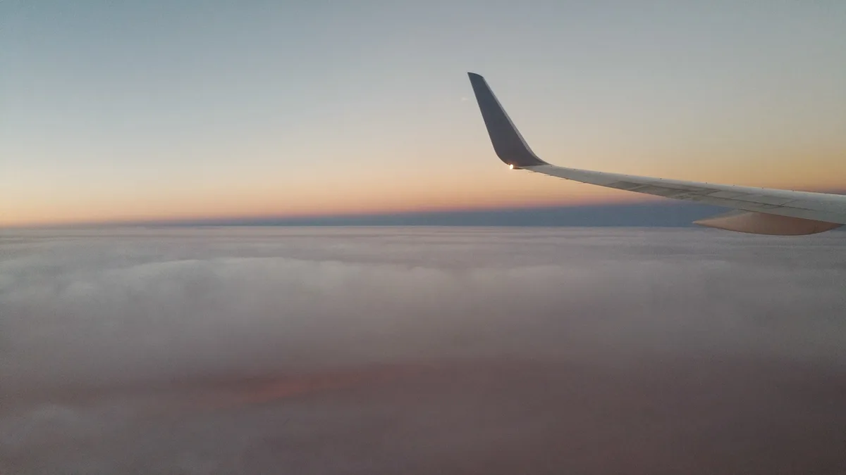 A serene view from an airplane window shows the wing against a twilight sky with soft orange and blue gradients above a sea…