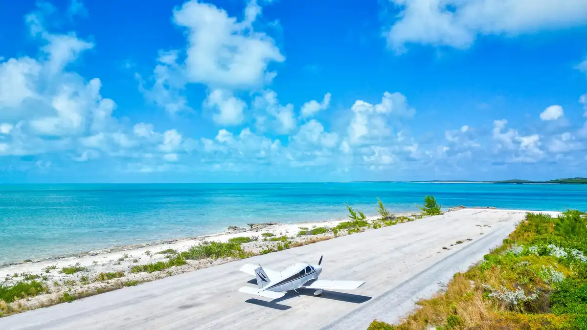 A small white single-engine airplane sits on a narrow paved runway next to a pristine white-sand beach with turquoise ocean…