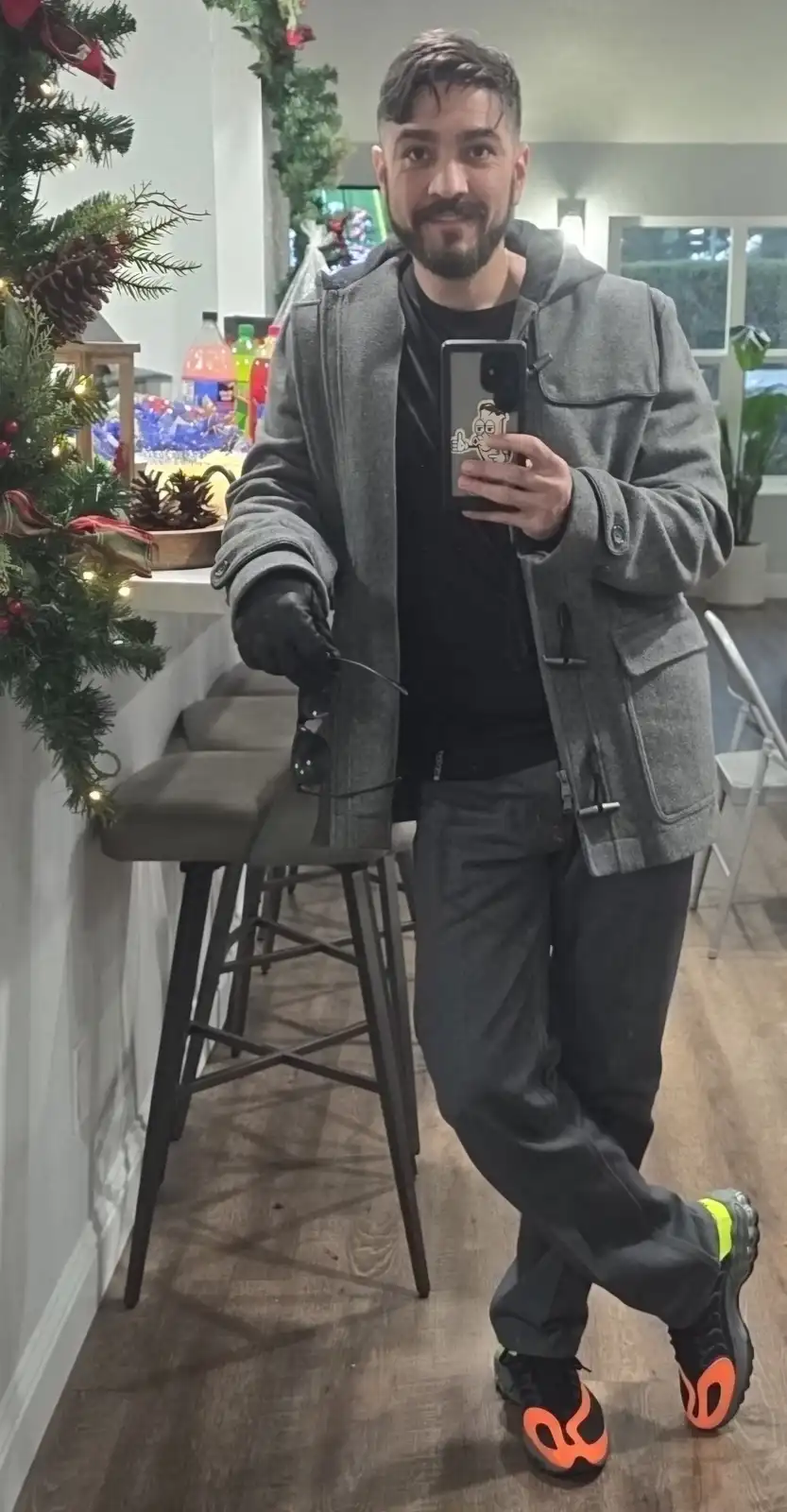 A man in a grey duffle coat, black gloves, and dark pants takes a mirror selfie while holding sunglasses. His bright orange…