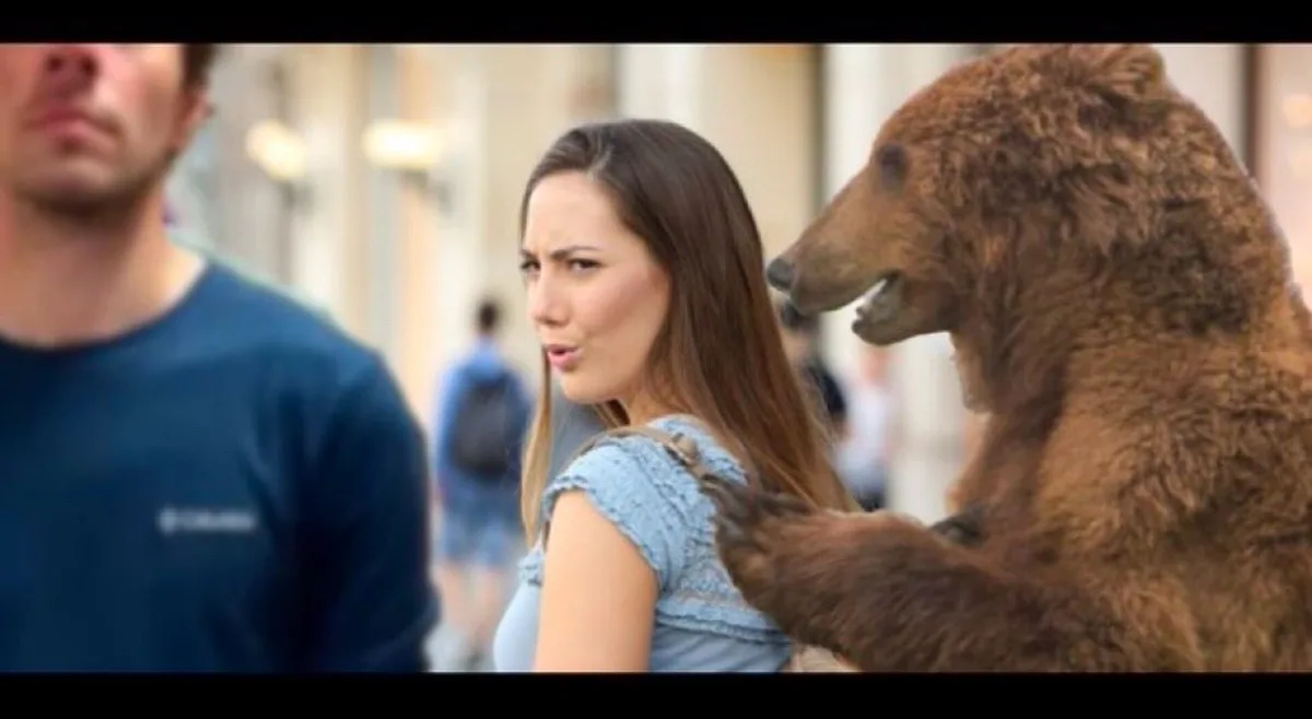 A woman in a blue top looks over her shoulder with a disgusted expression as a large brown bear stands behind her, its paw…
