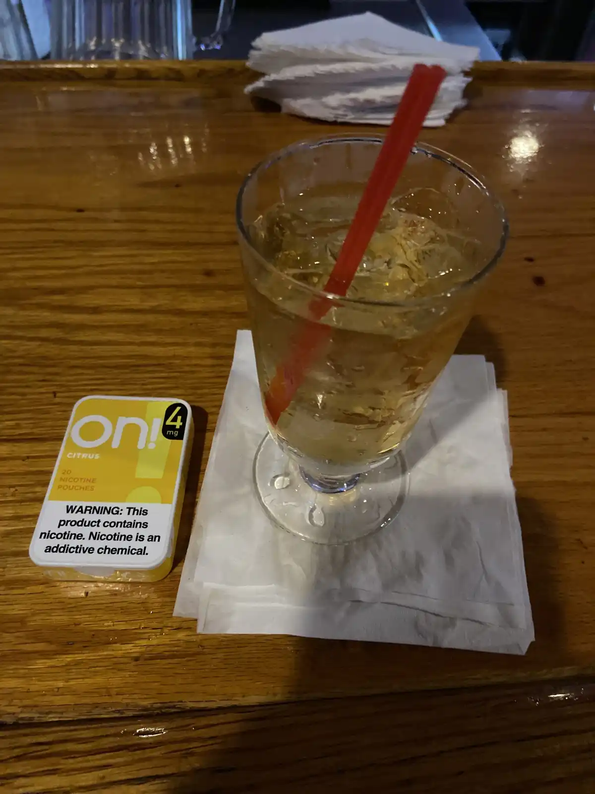 A close-up photo shows a glass of iced beverage with a red straw sitting on a white napkin on a wooden bar, next to a yellow…