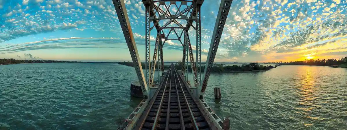A panoramic view from the center of a rusty metal truss bridge over water, looking down two parallel train tracks that…