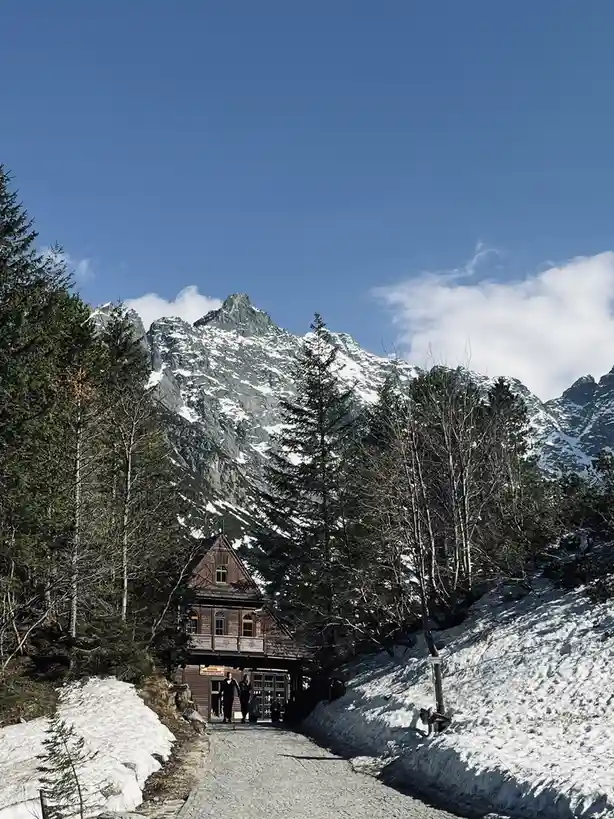 A scenic mountain landscape with snow-capped peaks under a clear blue sky, featuring a wooden building at the base of the…