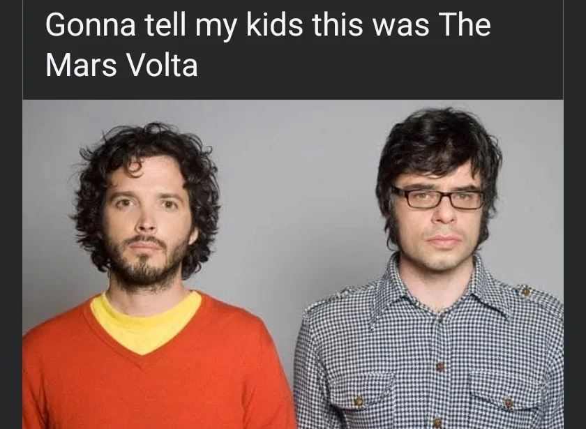 Two men, one with curly dark hair and a beard wearing an orange sweater over a yellow shirt, the other with glasses and dark…