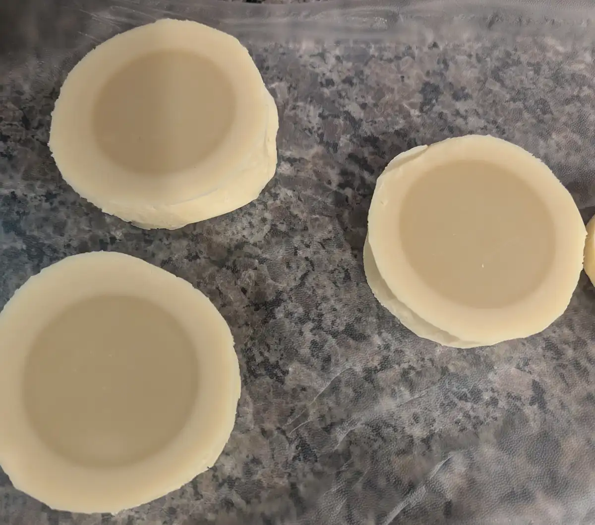 Three pale yellow, circular, cup-shaped cheese pieces sit on a gray speckled countertop, resembling empty eyes or bowls. The…