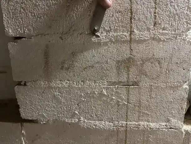 A close-up photo shows a hand holding a metal tool against a rough, light-colored brick wall. The wall has visible cracks…