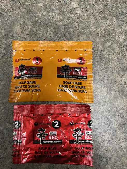 Two Nongshim Shin Ramyun Red Fiery Spicy Beef flavor soup base packets are laid out on a gray countertop. The top packet is…
