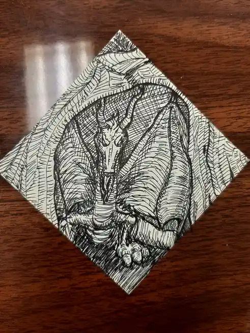 A black-and-white ink drawing on a diamond-shaped piece of paper, placed on a wooden surface, depicts a cat with a dragon's…