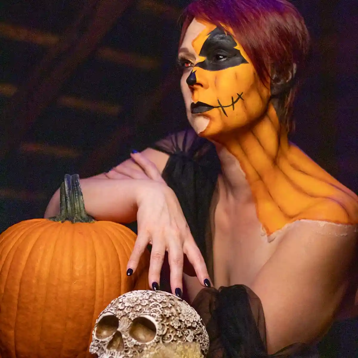 A person with Halloween-themed pumpkin face paint and orange body paint poses beside a real pumpkin and a decorative skull…