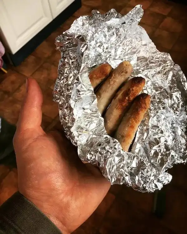 A hand holds a crumpled piece of aluminum foil containing four grilled sausages with browned, slightly charred exteriors…