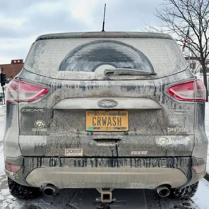 The rear of a dirty Ford Escape covered in mud and ice, with a yellow Iowa license plate reading 'CRWASH'. Stickers include…