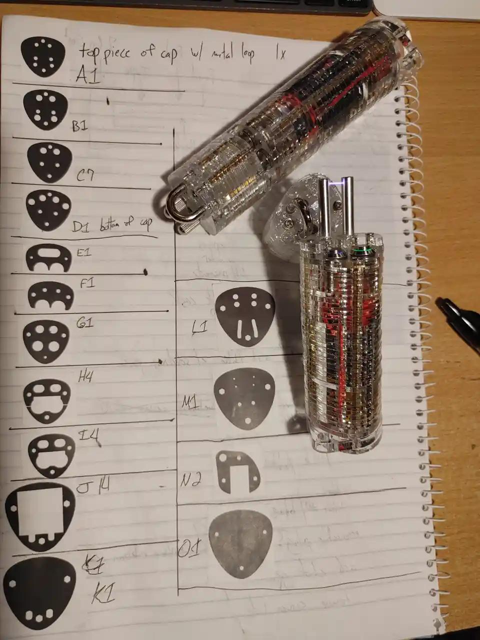 A notebook displays hand-drawn schematics of various guitar pick-shaped parts labeled A1 through K1, next to two…
