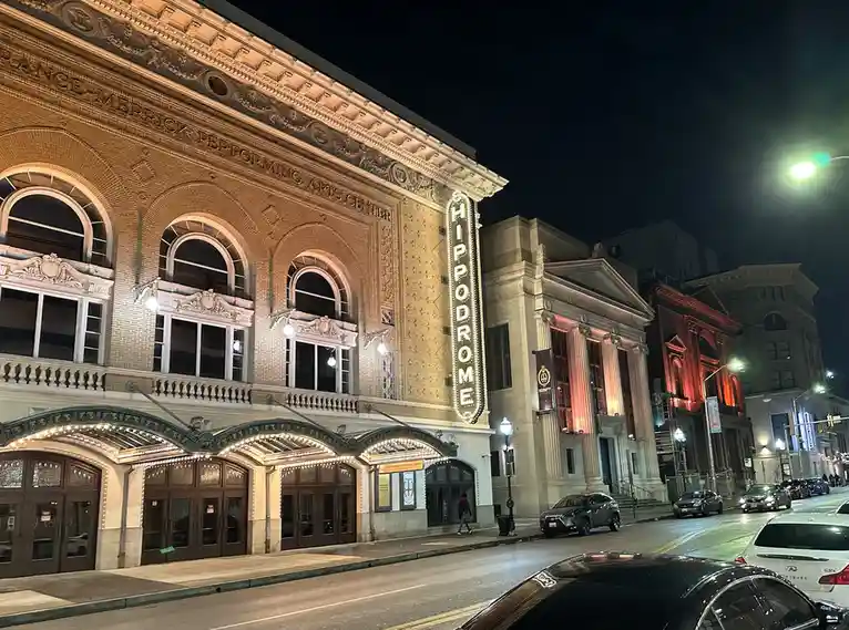 A nighttime photo of the ornate, illuminated facade of the Hippodrome Performing Arts Center, with its vertical marquee sign…