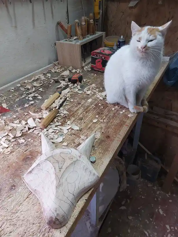A white and orange cat sits regally on a cluttered woodworking bench covered in sawdust, next to a partially carved wooden…