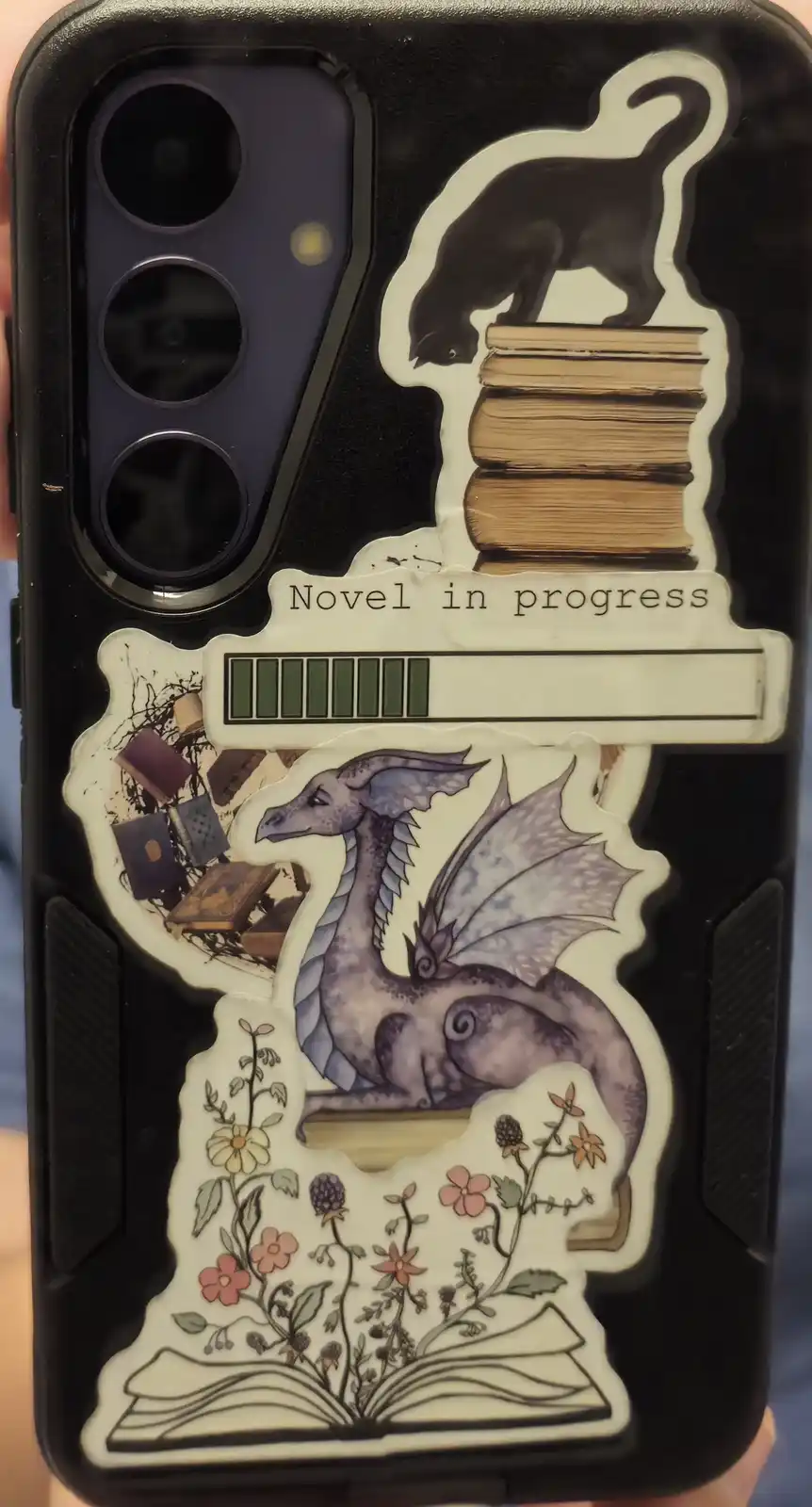A black phone case decorated with multiple stickers: a black cat perched atop a stack of books, a purple dragon resting…