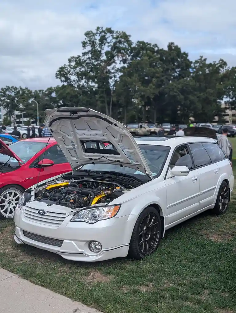 A white Subaru Legacy station wagon with its hood open, revealing a modified engine with prominent gold-colored exhaust…