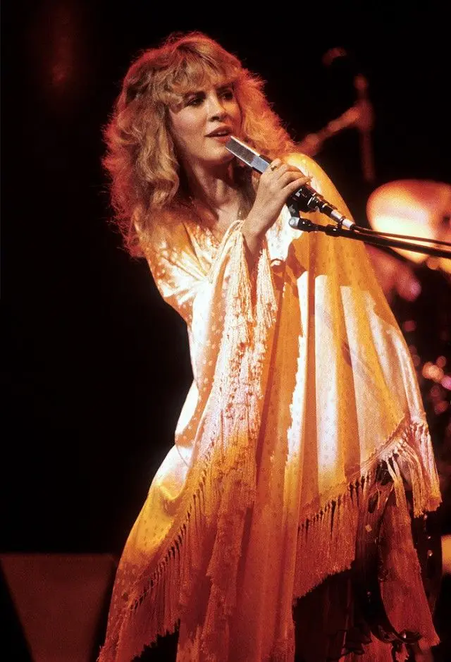 a woman with curly blonde hair wearing a flowing, fringed, orange-yellow robe sings into a microphone on a dimly lit stage…