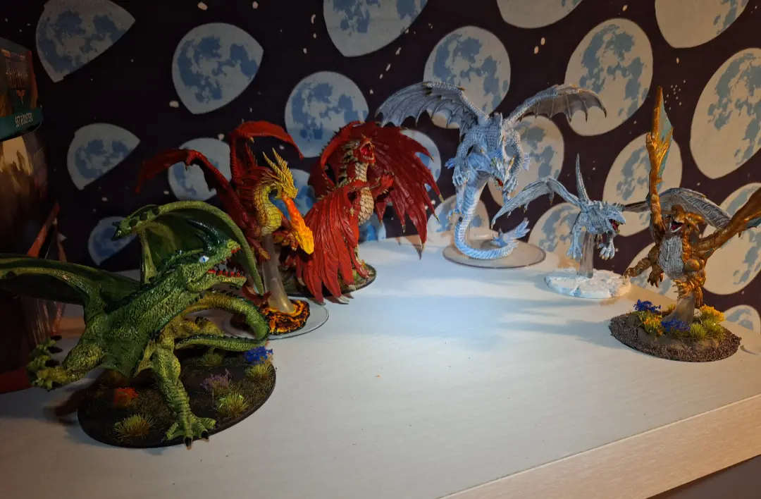 A collection of painted miniature dragons in various colors—green, red, blue, and gold—are arranged on a white shelf against…