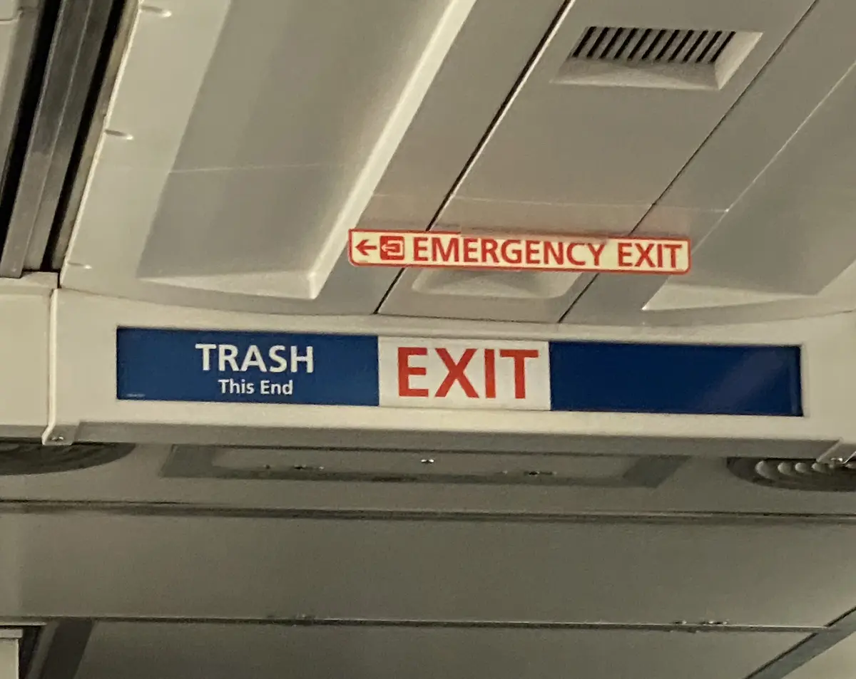 A close-up photo of airplane overhead signs shows a red 'EMERGENCY EXIT' sign with an arrow pointing left, directly above a…