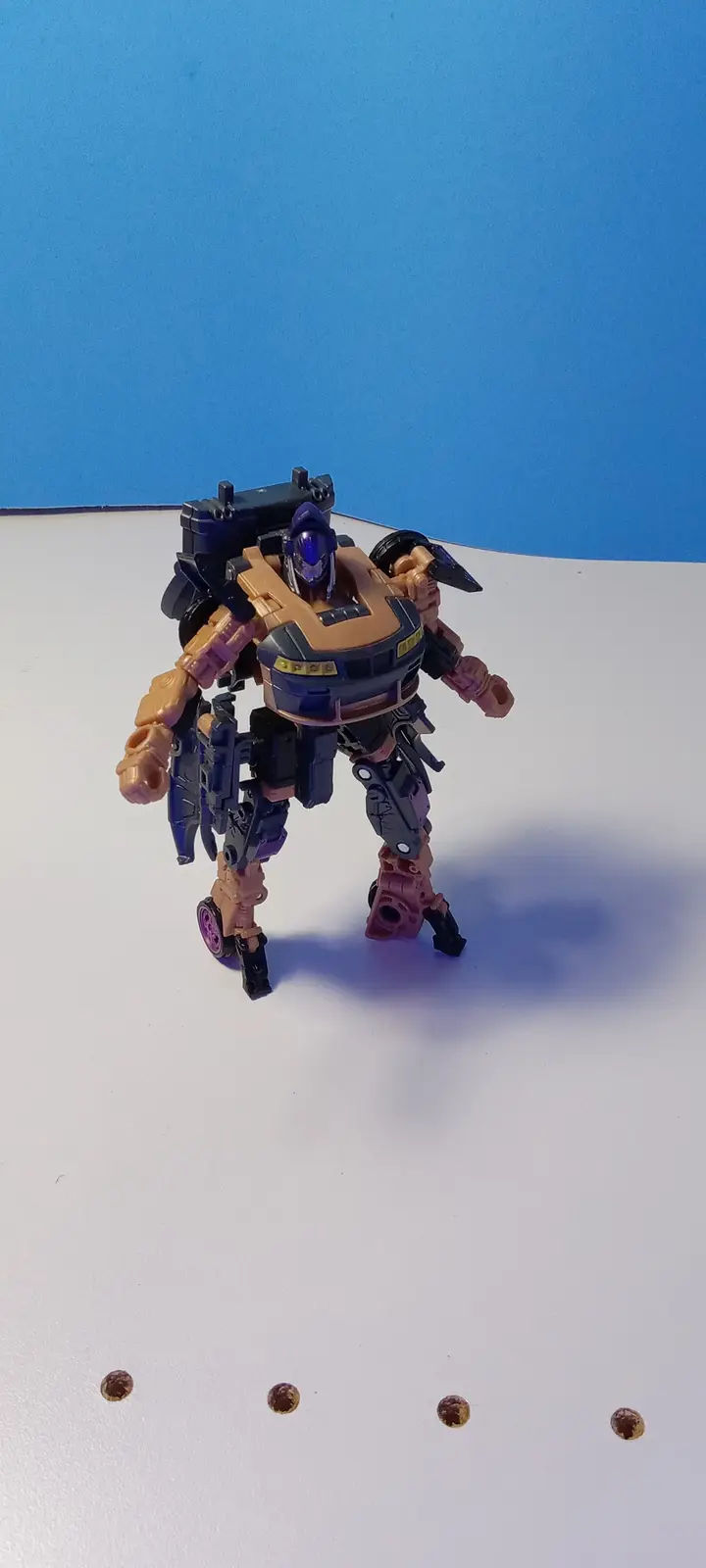 A tan and purple transformer toy stands on a white surface against a blue background, with four small brown nuts arranged in…