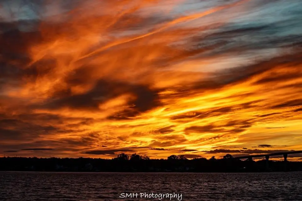 A dramatic sunset photograph showing fiery orange and yellow clouds over a body of water with a distant bridge and treeline…