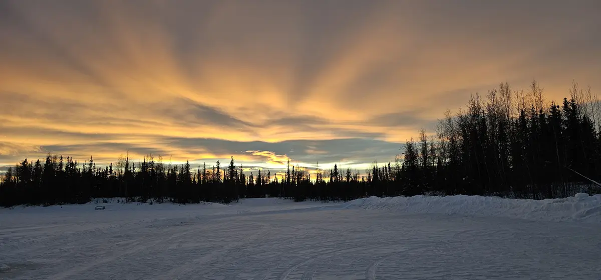 A wide winter landscape shows a snow-covered field with tracks, bordered by dark silhouetted trees against a dramatic sunset…