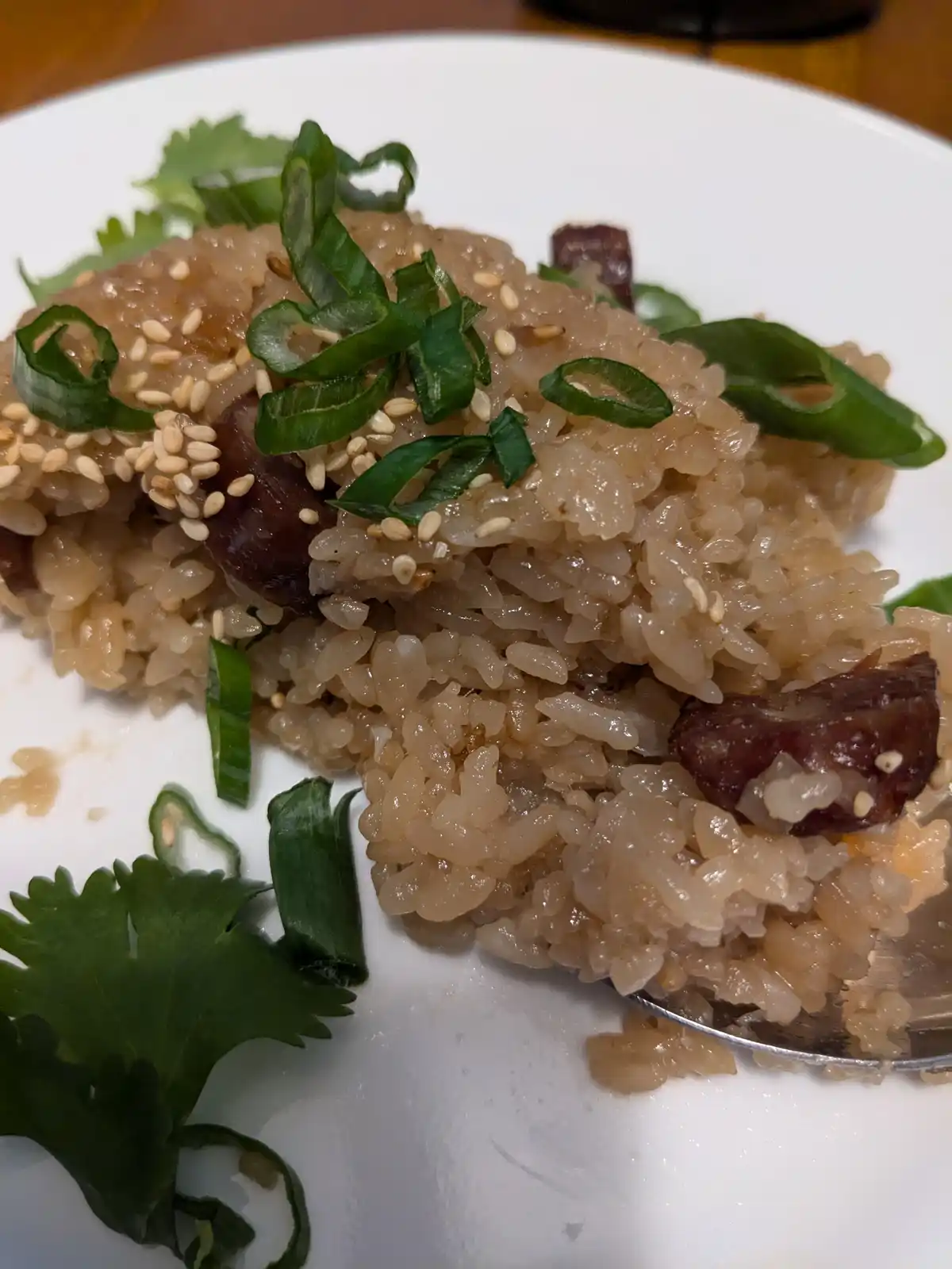 A close-up photo of a white plate holding a mound of brown rice mixed with dark meat chunks, topped with sesame seeds and…