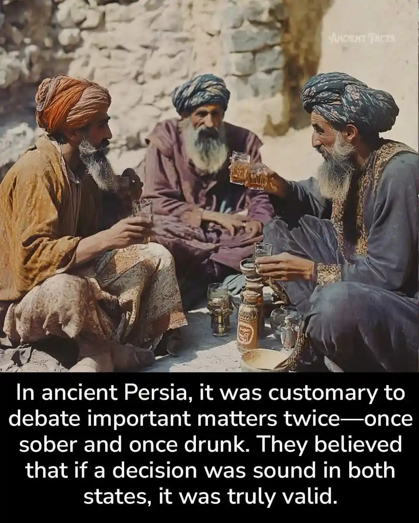 Three bearded men in traditional turbans sit on the ground against a stone wall, holding glasses of liquid, seemingly…
