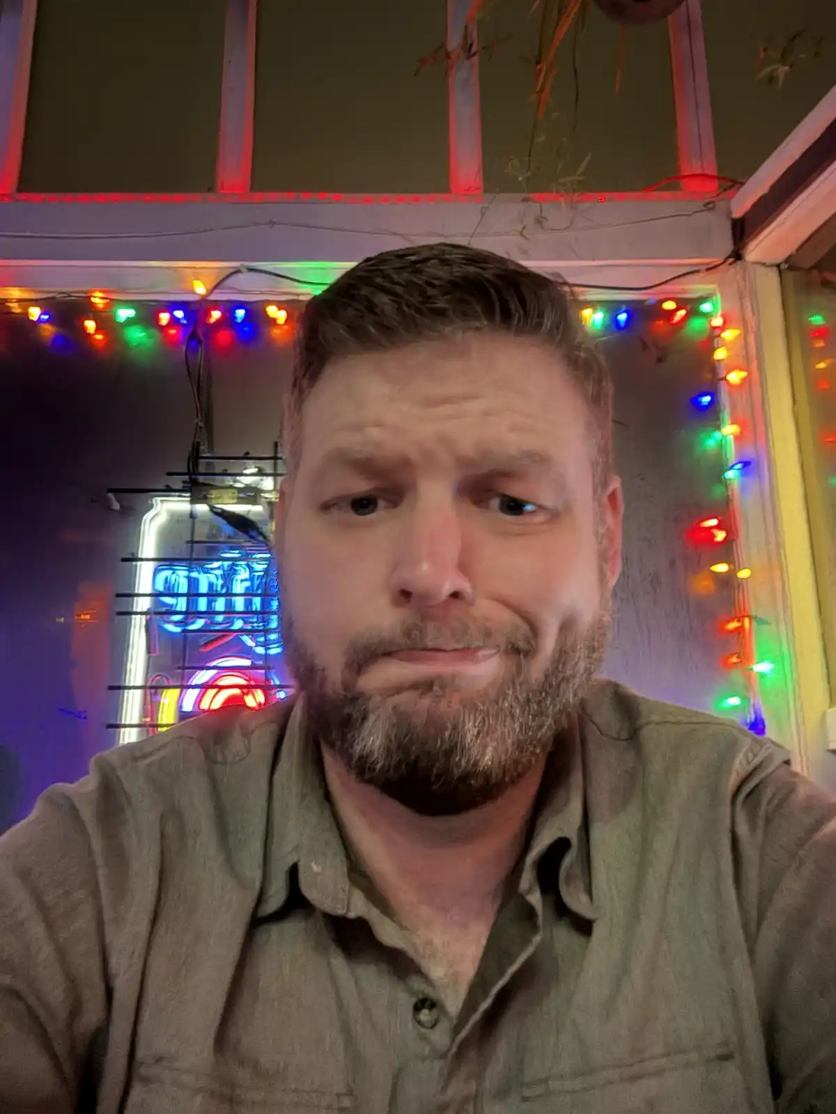 A man with a beard and short hair wearing a green button-up shirt looks directly at the camera with a slightly confused or…