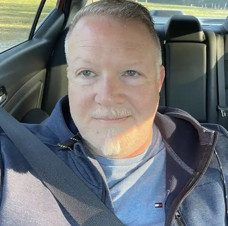 A man with gray hair and a goatee sits in a car, wearing a blue hoodie over a gray t-shirt with a Tommy Hilfiger logo…