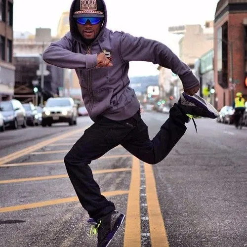 A man in a grey hoodie, black pants, and a baseball cap is mid-air on a city street, kicking one leg up while holding his…