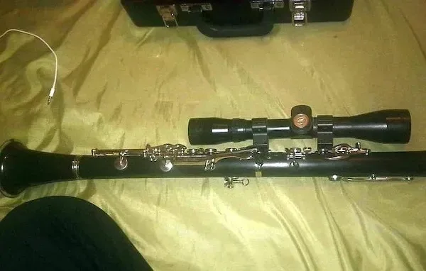 A clarinet lies horizontally on a bedspread, with a rifle scope mounted on top of it using brackets. In the background, a…