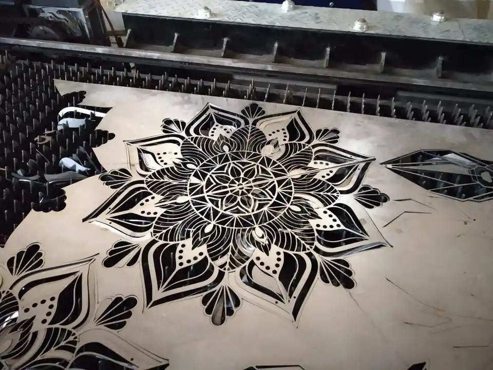 a close-up photo shows a large, intricate mandala pattern laser-cut into a sheet of metal, with the cut-out pieces revealing…